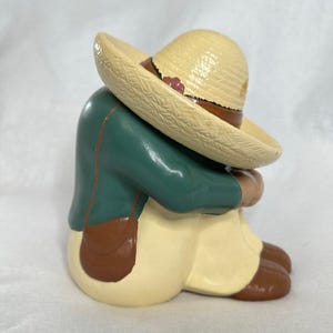 May include: A ceramic figurine of a person wearing a sombrero, with the head bowed. The figure is dressed in a green jacket, tan pants, and brown shoes. A brown satchel hangs from the shoulder. The sombrero is tan with a brown band and floral accents.