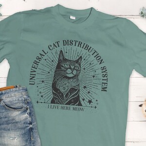 May include: A light green t-shirt with a black graphic of a cat with a starburst design. The text on the graphic reads "Universal Cat Distribution System I Live Here Meow".