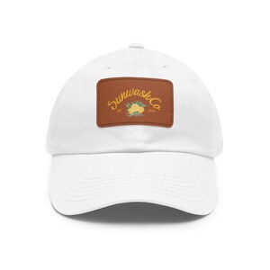 Puede incluir: Una gorra de béisbol blanca con un parche rectangular de cuero marrón en la parte delantera. El parche muestra el texto Sunwash Co en letra cursiva amarilla, una ilustración de cítricos amarillos y hojas verdes, y el texto EST 2018.