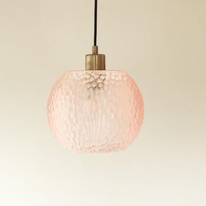 Könnte beinhalten: Eine rosa, strukturierte Glas-Pendelleuchte mit rundem Schirm. Die Leuchte hat eine messingfarbene Oberseite und ein schwarzes Kabel. Der Schirm hat eine strukturierte Oberfläche, und das Licht hängt vor einem neutralen Hintergrund.