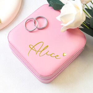 Op de afbeelding: Een roze sieradendoosje met de naam "Alice" in gouden letters, een klein gouden hartje en twee zilveren ringen. Een gouden ketting met de naam "Linda" staat op de achtergrond. Witte rozen en groene bladeren zijn op de achtergrond.