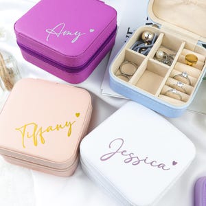 Peut inclure: Plusieurs boîtes à bijoux carrées personnalisées, de couleurs violet, rose et blanche. Chaque boîte porte un nom écrit en script. Une boîte bleue est ouverte, révélant des bagues et autres bijoux.