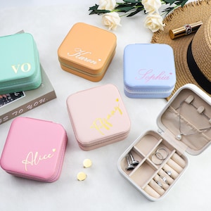 May include: Several pastel-coloured jewellery boxes, including mint green, peach, light blue, and pink, each with a name in a cursive font. One box is open, displaying compartments for jewellery storage. The boxes are square-shaped.