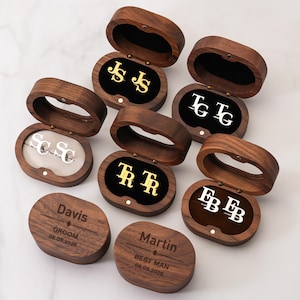 Puede incluir: Cajas ovaladas de madera con gemelos personalizados. Las cajas abiertas revelan gemelos dorados y plateados con iniciales. Dos cajas cerradas están grabadas con nombres y fechas. Un regalo ideal para los padrinos.