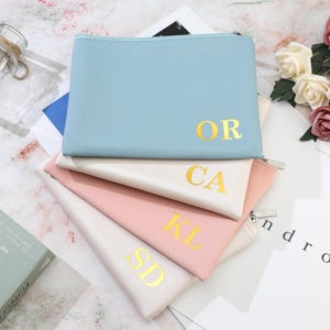 May include: Four pastel-coloured zippered pouches stacked on top of each other. The top pouch is light blue with gold lettering "OR". The pouches below are cream, pink, and cream, each with gold initials. A glass bottle and flowers are in the background.