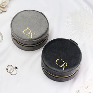 May include: Two round, grey and black velvet jewellery cases with gold zips and initial monograms. A gold and silver ring set and a pearl necklace are also visible. The cases are on a white surface with a feather and flowers.