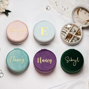 Könnte beinhalten: Sechs runde Schmuckkästchen aus Samt in Pastellfarben, darunter Rosa, Blau, Türkis, Lila und Grün. Jede Box hat einen goldenen Namen oder Initialen auf dem Deckel. Eine Box ist geöffnet und zeigt Fächer für Ringe und Halsketten. Eine stilvolle Aufbewahrungslösung.