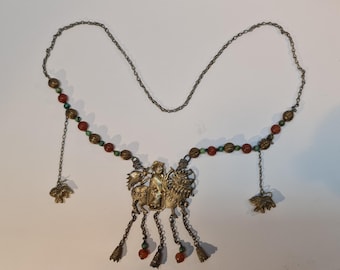 Op de afbeelding: Een zilveren ketting met een hanger met een figuur die een mythisch beest berijdt. De hanger is versierd met rode en groene kralen. De ketting heeft meerdere hangende bedels.