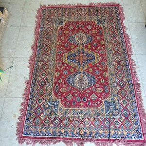 May include: A rectangular red rug with an intricate geometric pattern in blue, yellow, and white. The rug has a central medallion design and a fringed border. The rug's dimensions are not visible in the image.