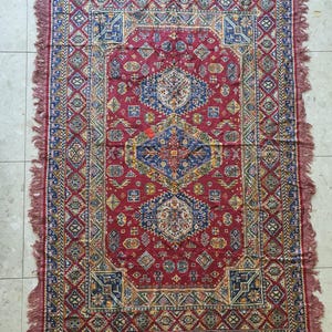 May include: A rectangular, red rug with an intricate geometric pattern. The rug features a central medallion design in blue, yellow, and white, surrounded by a border of repeating shapes. The edges are finished with a fringe.