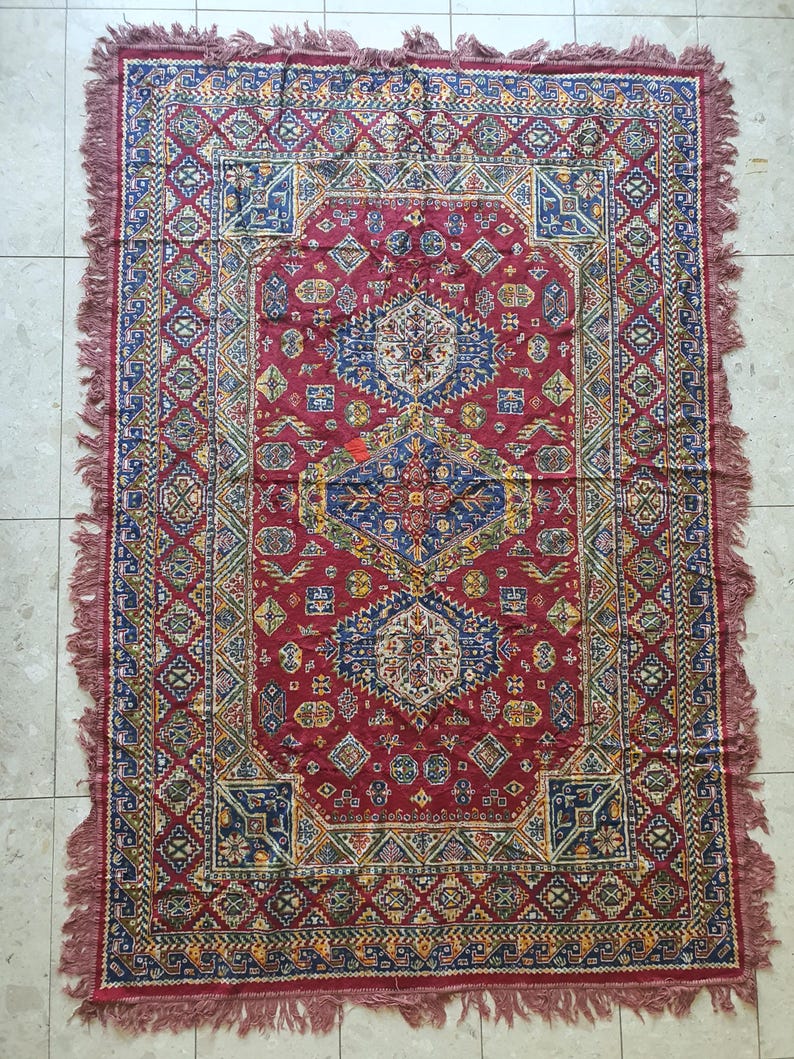 May include: A rectangular, red rug with an intricate geometric pattern. The rug features a central medallion design in blue, yellow, and white, surrounded by a border of repeating shapes. The edges are finished with a fringe.