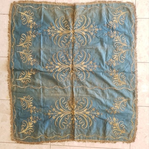 Puede incluir: Textil cuadrado azul con bordados dorados. El bordado presenta diseños florales y de desplazamiento dispuestos simétricamente. Los bordes están acabados con flecos dorados. El textil parece ser vintage o antiguo.