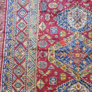 May include: Close-up of a section of a red, patterned rug. The rug features geometric designs in blue, green, yellow, and white. The edges have a fringe. The rug's intricate patterns and rich colors suggest a traditional or ethnic style.