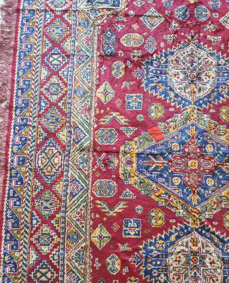 May include: Close-up of a section of a red, patterned rug. The rug features geometric designs in blue, green, yellow, and white. The edges have a fringe. The rug's intricate patterns and rich colors suggest a traditional or ethnic style.