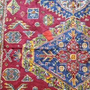May include: Close-up of a section of a red, blue, and yellow patterned rug. The rug features geometric shapes and floral motifs. The colors are vibrant, with a mix of deep reds, blues, and yellows. The rug appears to be made of a soft, textured material.