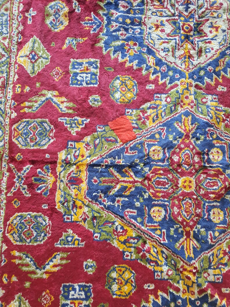 May include: Close-up of a section of a red, blue, and yellow patterned rug. The rug features geometric shapes and floral motifs. The colors are vibrant, with a mix of deep reds, blues, and yellows. The rug appears to be made of a soft, textured material.