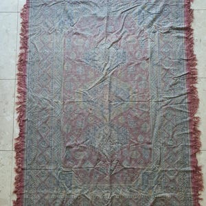 May include: A rectangular, patterned textile with a faded red and blue color scheme. The textile features an intricate design with a border and fringe along the edges. The textile is likely a rug or blanket.