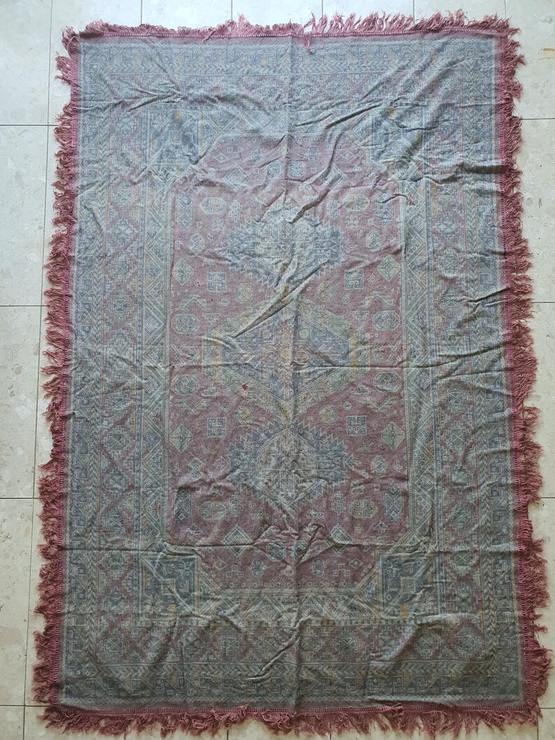 May include: A rectangular, patterned textile with a faded red and blue color scheme. The textile features an intricate design with a border and fringe along the edges. The textile is likely a rug or blanket.