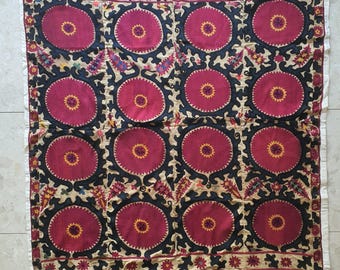 May include: A square, patterned textile featuring a repeating design of large, circular motifs in shades of red and black. The circles have yellow centers and are set against a beige background with floral accents. The border is white.