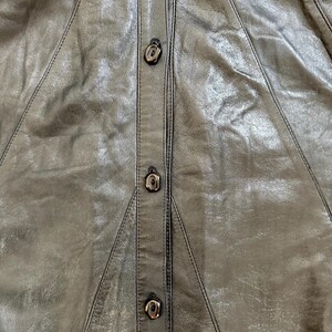 May include: Close-up of a black leather garment with three decorative buttons down the center. The leather has a slightly textured appearance with visible stitching details. The buttons are a dark, metallic color.