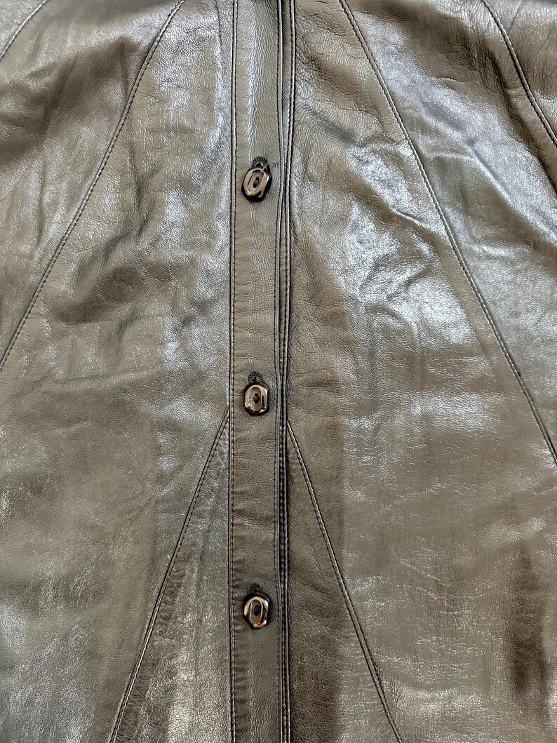 May include: Close-up of a black leather garment with three decorative buttons down the center. The leather has a slightly textured appearance with visible stitching details. The buttons are a dark, metallic color.