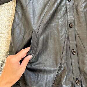 May include: Black leather coat with a button-up front and a tailored design. The coat features a classic collar, pockets, and a knee-length cut. The leather appears to be soft and supple, with visible stitching details. A timeless outerwear piece.