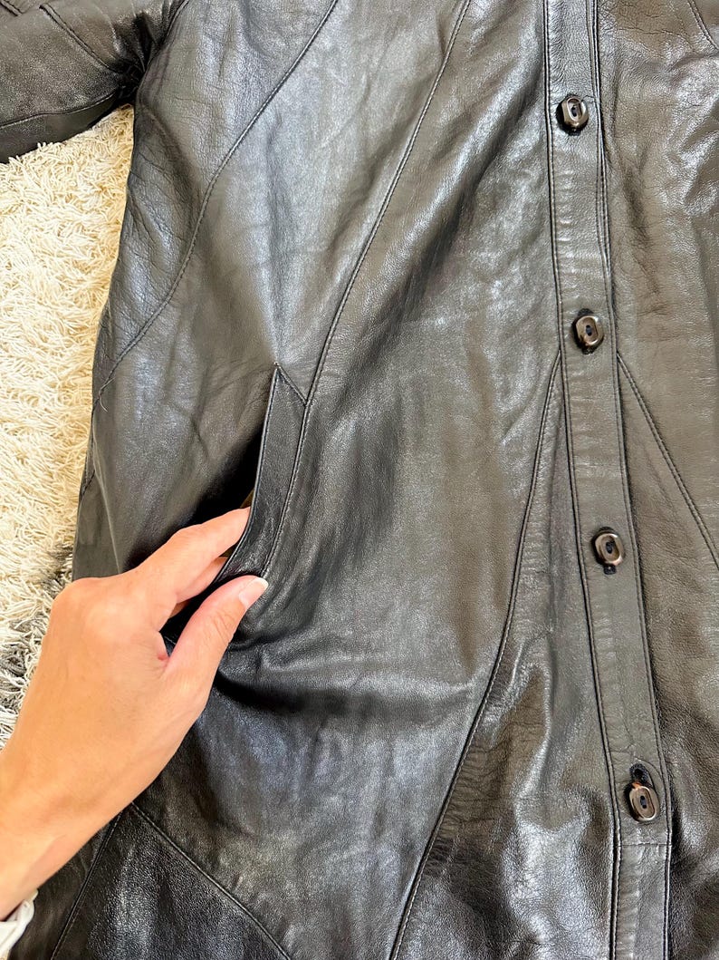 May include: Black leather coat with a button-up front and a tailored design. The coat features a classic collar, pockets, and a knee-length cut. The leather appears to be soft and supple, with visible stitching details. A timeless outerwear piece.