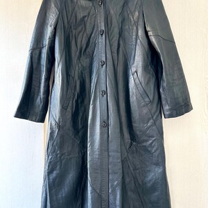 May include: A long, black leather coat with a button-up front and a collared neckline. The coat has a tailored fit, with seams creating a panel design. The interior lining is a patterned fabric. The coat is hanging on a wooden hanger.