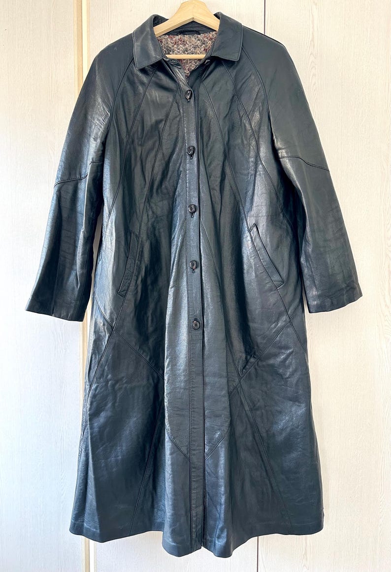 May include: A long, black leather coat with a button-up front and a collared neckline. The coat has a tailored fit, with seams creating a panel design. The interior lining is a patterned fabric. The coat is hanging on a wooden hanger.