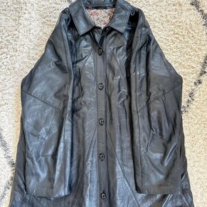 May include: A long, black leather coat with a button-up front and a collared neckline. The coat has a patchwork design and a floral patterned lining visible at the collar. The coat is laid flat on a textured surface.