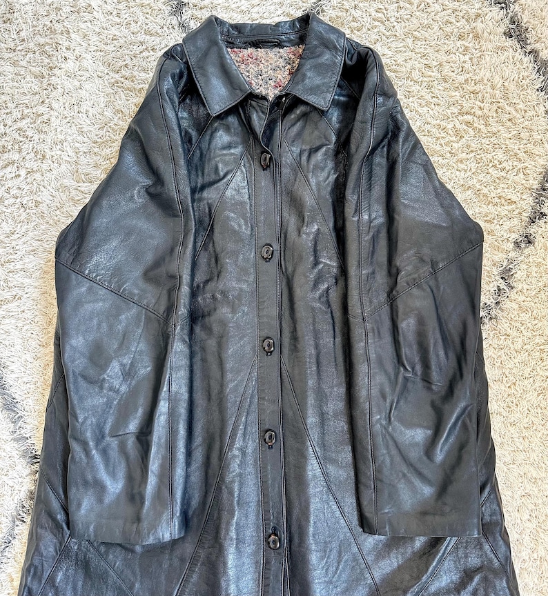 May include: A long, black leather coat with a button-up front and a collared neckline. The coat has a patchwork design and a floral patterned lining visible at the collar. The coat is laid flat on a textured surface.