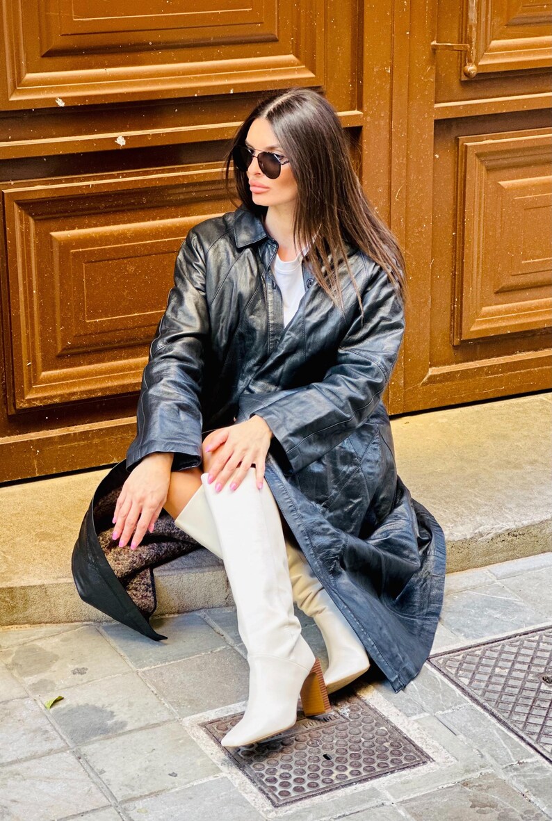 May include: A black leather trench coat with a quilted design. The coat is worn with white knee-high boots with a block heel. The person is wearing sunglasses and sitting on a stone step in front of a brown wooden door.