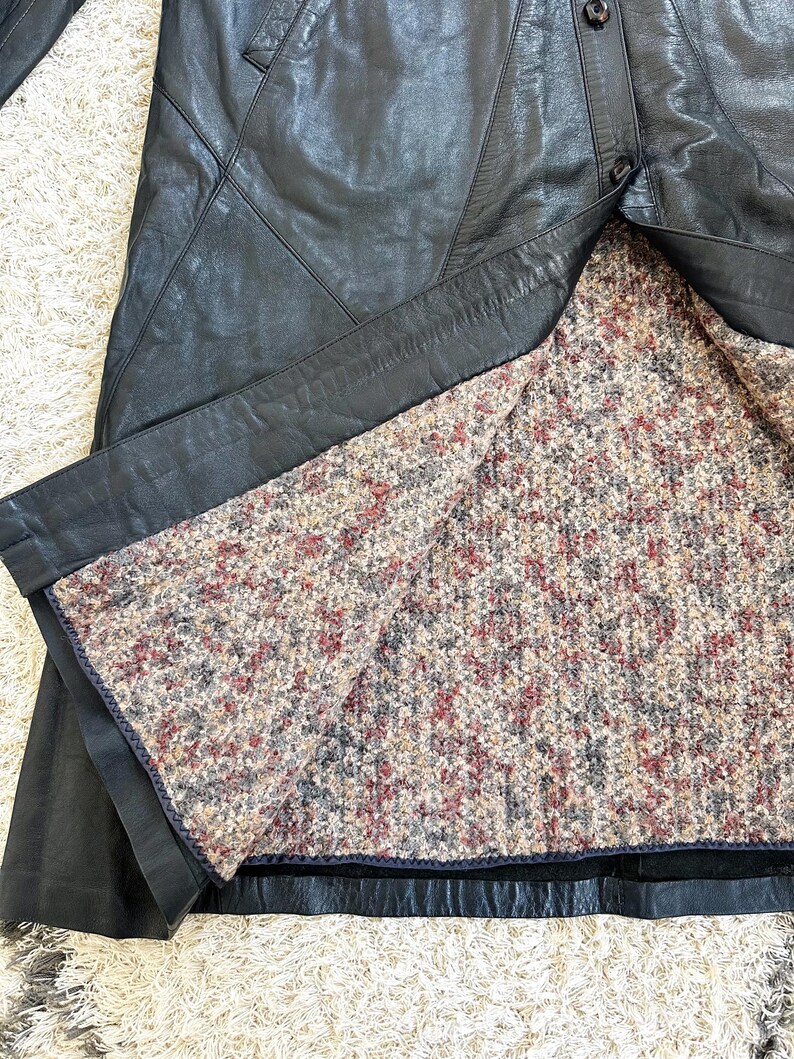 May include: Black leather coat with a textured, multi-colored interior lining. The coat features a button closure and a classic design. The lining has a pattern of red, brown, and gray flecks.