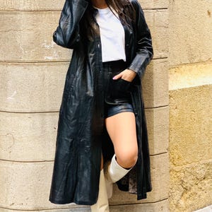 May include: A long black leather coat with a button-down front, paired with black shorts and white knee-high boots. The outfit includes a white t-shirt. The model is leaning against a textured stone wall.