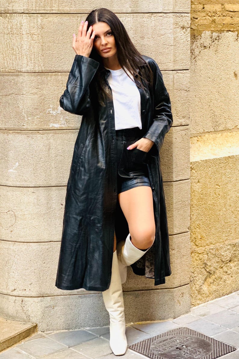 May include: A long black leather coat with a button-down front, paired with black shorts and white knee-high boots. The outfit includes a white t-shirt. The model is leaning against a textured stone wall.