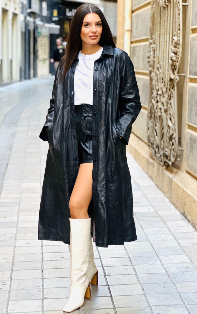 May include: A long black leather coat with a button-down front, worn over a white t-shirt and black shorts. The outfit is completed with cream-colored knee-high boots with a block heel. The model is standing on a cobblestone street.