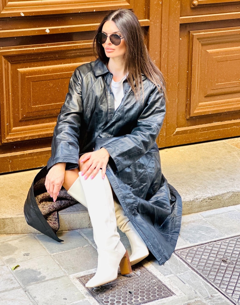 May include: A black leather trench coat with a classic collar and button closure. The coat is paired with white knee-high boots with a block heel. The person is wearing sunglasses.