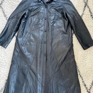 May include: A long, black leather coat with a button-up front and a collared neckline. The coat features a tailored design with geometric paneling and two front pockets. The interior lining is patterned. The coat is displayed on a neutral-colored rug.