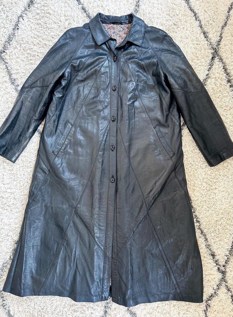 May include: A long, black leather coat with a button-up front and a collared neckline. The coat features a tailored design with geometric paneling and two front pockets. The interior lining is patterned. The coat is displayed on a neutral-colored rug.