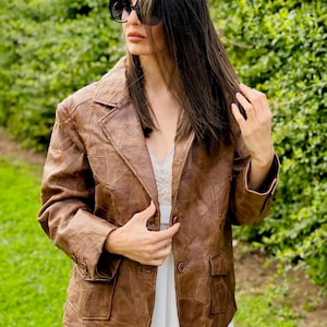 Può includere: Una giacca di pelle marrone con un design testurizzato. La giacca ha una chiusura con bottoni e due tasche. La modella indossa una gonna bianca e occhiali da sole.