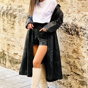 May include: A woman wearing a white t-shirt, black leather shorts, a long black leather coat, and cream-colored knee-high boots with a block heel. The outfit is set against a textured stone wall and a paved walkway.