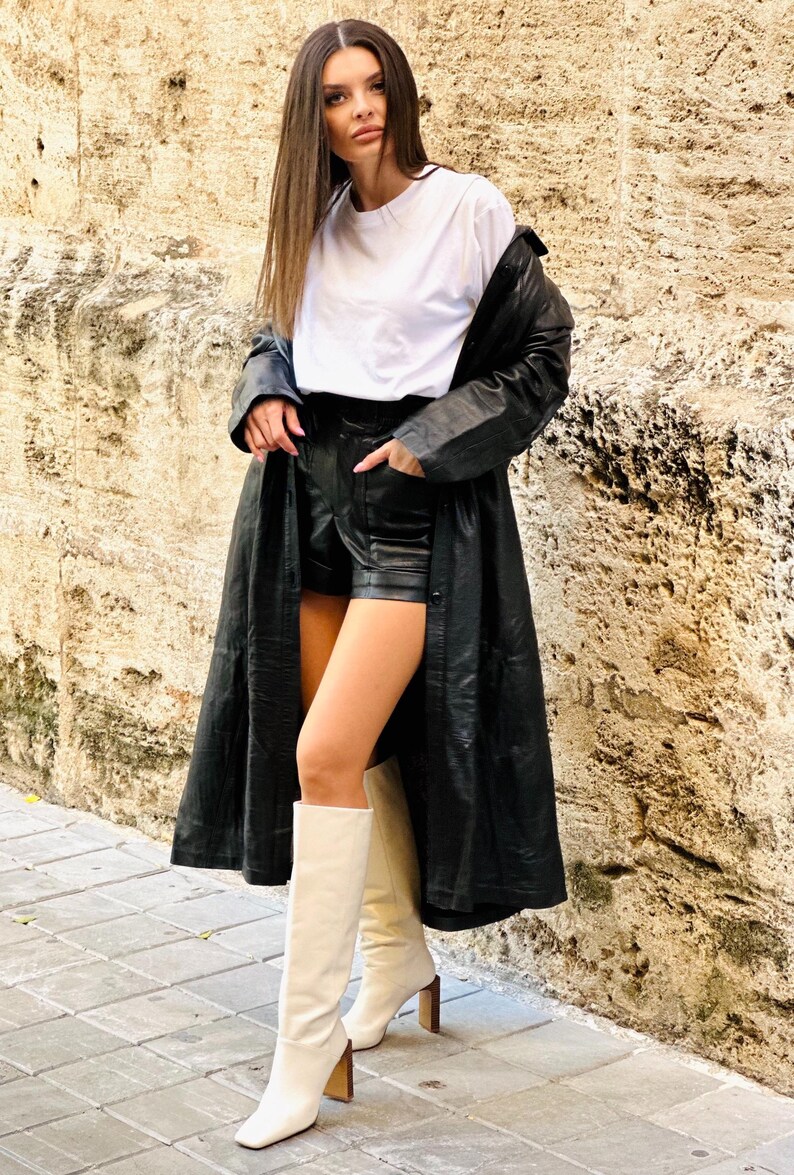 May include: A woman wearing a white t-shirt, black leather shorts, a long black leather coat, and cream-colored knee-high boots with a block heel. The outfit is set against a textured stone wall and a paved walkway.