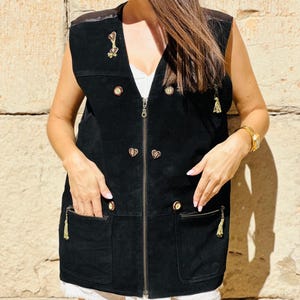 Op de afbeelding: Zwarte suède vest met een V-hals, ritssluiting en goudkleurige knopen. Het vest heeft twee voorzakken met gouden kwastjes en een decoratieve broche. Het vest wordt over een witte top gedragen.