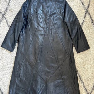May include: Black leather coat with a collared neckline and long sleeves. The coat features a geometric design on the back with stitched panels. The coat is knee-length and appears to be vintage.