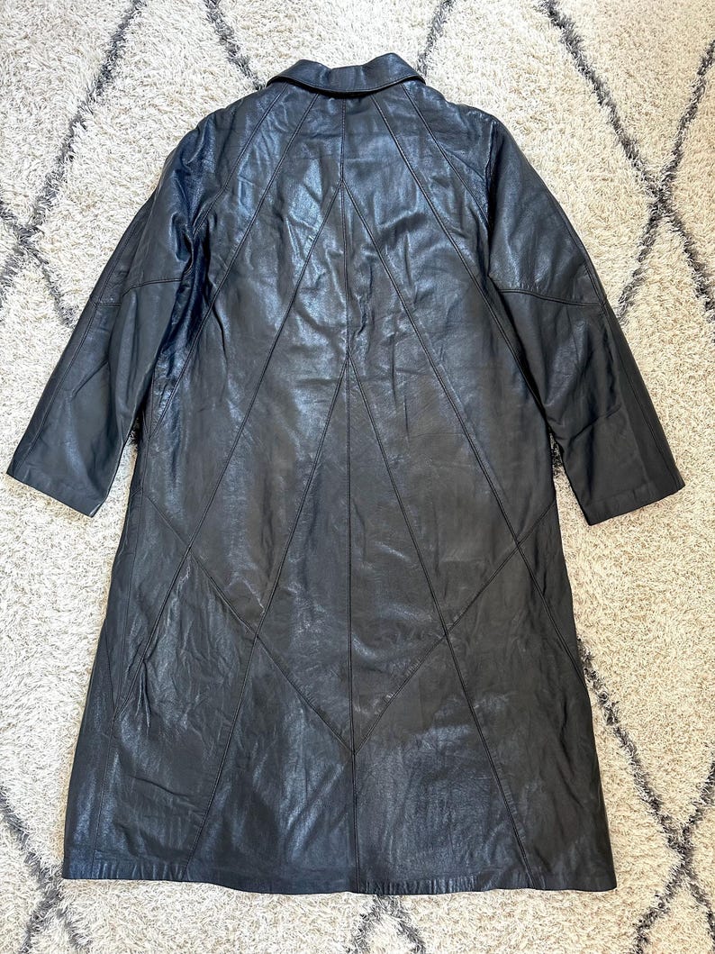 May include: Black leather coat with a collared neckline and long sleeves. The coat features a geometric design on the back with stitched panels. The coat is knee-length and appears to be vintage.