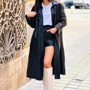 May include: A long black leather coat, black shorts, a white t-shirt, and cream-colored knee-high boots. The outfit is worn outdoors on a sunny day. The coat is buttoned up and reaches the ankles.