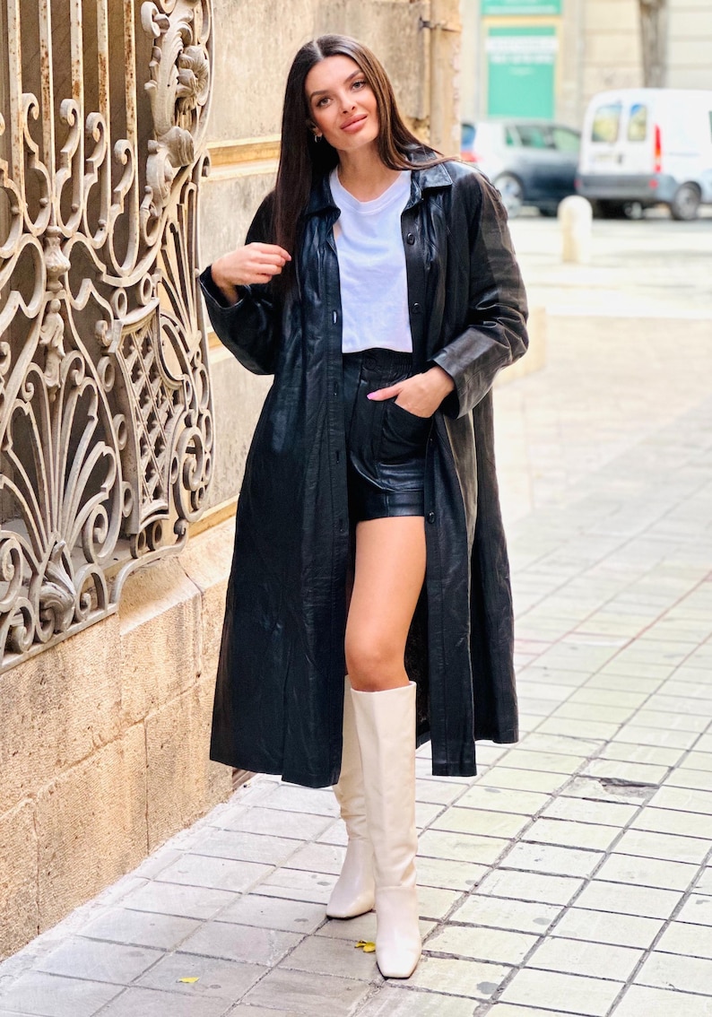 May include: A long black leather coat, black shorts, a white t-shirt, and cream-colored knee-high boots. The outfit is worn outdoors on a sunny day. The coat is buttoned up and reaches the ankles.