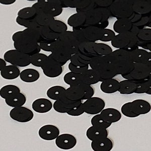 May include: A large quantity of black round sequins, scattered on a white surface. The sequins are flat and have a hole in the center.