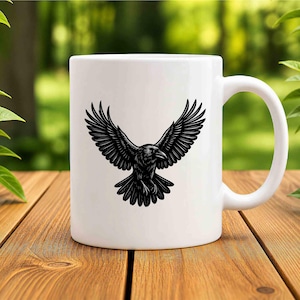 May include: A white ceramic mug featuring a black graphic illustration of a raven with its wings spread wide in flight. The bird is depicted in a detailed, etched style centered on the front of the mug, which has a smooth finish and a handle.