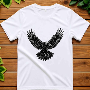 May include: A white short-sleeved t-shirt featuring a centered black graphic illustration of a raven with its wings spread wide in flight. The bird is depicted with detailed feather textures and a sharp beak, positioned against the plain white fabric.